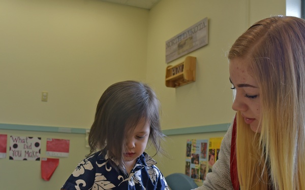 A young child and an older female (likely a teacher or aide) are engaged in a hands-on play-dough activity at a table in what appears to be a preschool or early childhood classroom. The adult is leaning in to assist the child, who is wearing a blue floral shirt and working with small rolling tools and stamps. The classroom setting includes educational posters, decorations, and learning materials visible in the background.