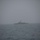 A military naval vessel is visible at a distance on open water under heavily overcast, foggy conditions. The ship appears to be a patrol or corvette-class warship, identifiable by its superstructure, mast, and radar equipment. The image is taken from another vessel or elevated vantage point across choppy gray seas.