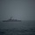 A military naval vessel is photographed at sea under heavily overcast, foggy conditions. The ship appears to be a small-to-medium sized warship, likely a patrol or coastal defense vessel, sitting stationary or moving slowly on moderately choppy open water. The dark, muted tones and low visibility create a somber, austere atmosphere.
