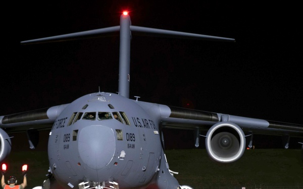 A U.S. Air Force C-17 Globemaster III (tail number 0189, 164th Airlift Wing) is being marshaled on a tarmac at night by a ground crew member wearing a reflective orange vest and holding illuminated red wands. The aircraft is facing forward with all four engines visible and navigation/anti-collision lights active. The scene is lit primarily by the aircraft's own lighting and ambient airfield illumination.