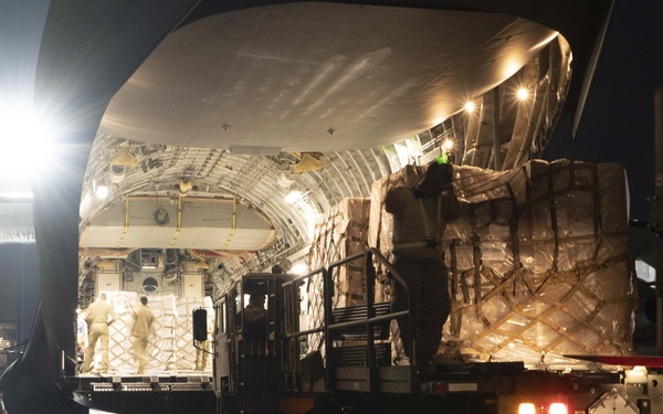 A large military or cargo aircraft is being loaded with palletized freight during nighttime operations on a wet tarmac. Ground crew personnel and specialized loading equipment are actively transferring strapped cargo pallets into the aircraft's open rear cargo bay. The scene is illuminated by aircraft and ground support lighting against a dark sky.