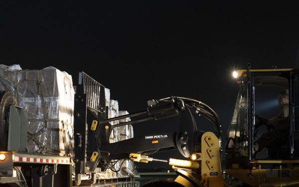 A nighttime loading operation showing a yellow forklift/loader engaging with cargo pallets on a flatbed trailer. The cargo appears to be military or industrial supplies wrapped in plastic and secured with straps. The scene is illuminated by vehicle lights and a nearby overhead light.