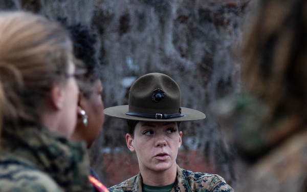 A female U.S. Marine drill instructor wearing the distinctive campaign cover (drill sergeant hat) is speaking with a group of individuals, some in military camouflage uniforms. The setting appears to be outdoors with trees visible in the background. The interaction suggests a briefing, orientation, or instructional session.