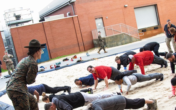 A female Marine drill instructor in camouflage uniform and campaign cover is overseeing a group of civilians performing push-ups in a sand pit, likely during a military orientation or 'citizen warrior' type event. The participants are wearing casual civilian clothing and some have lanyards/badges, suggesting they are visitors or media personnel experiencing military training. The setting appears to be a military installation with brick buildings visible in the background.