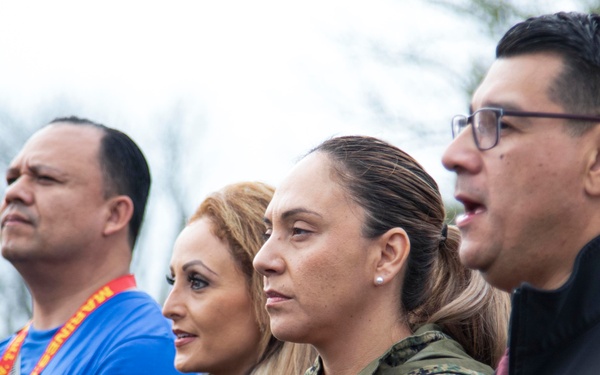 A group of adults standing together outdoors, appearing to observe or attend an event. Several individuals are wearing lanyards with red and yellow coloring, and one person is wearing military camouflage attire. The setting appears to be an outdoor gathering, possibly a military or community event.