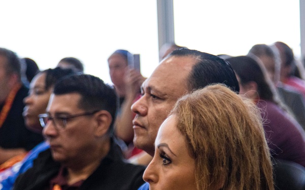 A group of adults are seated in rows, appearing to attend a presentation, conference, or meeting in a well-lit indoor venue. The foreground shows three individuals in sharp focus, with additional attendees visible in the background. Several participants are wearing lanyards, suggesting a formal or organized event.