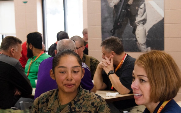A group of people are gathered in what appears to be a military dining facility or meeting room. A female service member in US Marine Corps digital camouflage (MARPAT) uniform is engaged in conversation with a civilian woman wearing a navy blue shirt and a lanyard. The setting suggests an organized event, possibly a family day, orientation, or community outreach program.