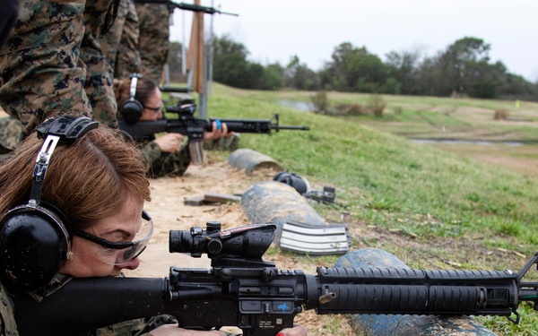 Multiple individuals in U.S. Marine Corps MARPAT camouflage uniforms are participating in a rifle qualification or familiarization exercise at an outdoor shooting range. The foreground shooter, a woman in prone position, is aiming an M16A4 or M4-style rifle equipped with an ACOG optic over a log barrier. Spent brass casings are visible on the ground, indicating active firing.