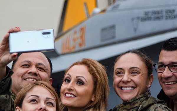A group of five people, four wearing military camouflage uniforms and one in civilian clothing, are posing for a selfie in front of a military aircraft. The setting appears to be a military base or air show, with a U.S. Marine Corps or Navy aircraft visible in the background. The group appears cheerful and relaxed, smiling broadly for the photo.