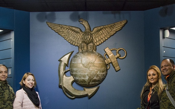 Four visitors pose in front of a large U.S. Marine Corps Eagle, Globe, and Anchor emblem at what appears to be a Marine Corps museum. A quote from President Ronald Reagan (1985) is inscribed on the blue wall below the emblem. Display cases on either side contain historical Marine Corps uniforms on mannequins.