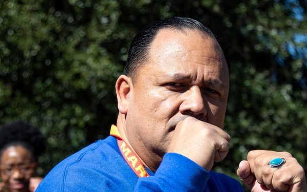 A man in a blue sweatshirt is posed in a boxing or fighting stance with both fists raised, one near his face. He is wearing a red and yellow lanyard with partial text visible reading 'MAR'. A woman in a black jacket with a lanyard is visible in the background, appearing to mimic a similar stance.