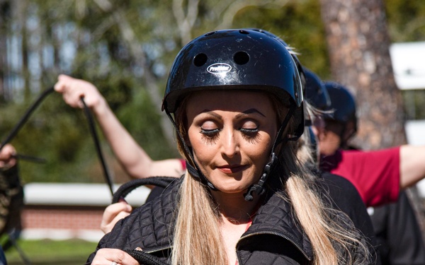 A woman wearing a black safety helmet and quilted jacket is handling black ropes or harness lines, appearing to be preparing for or participating in an outdoor adventure activity. She is focused downward on the equipment in her hands. Several other participants are visible in the background, also holding ropes, suggesting a group activity or training session.