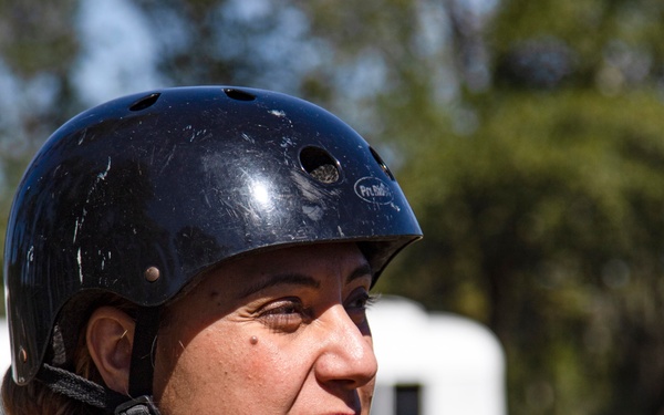 A woman wearing a black protective helmet and digital camouflage military uniform is photographed in a three-quarter profile view outdoors. She appears to be in a military training or operational environment, with a white vehicle or trailer visible in the blurred background. The image is a close-up portrait taken in natural daylight.
