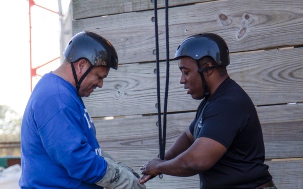Two individuals are standing near a large wooden wall structure in an outdoor setting. A person in military camouflage uniform is assisting a civilian in a blue sweatshirt with rappelling or climbing equipment. Both are wearing protective helmets, and the civilian is wearing work gloves while being fitted with a harness and rope system.