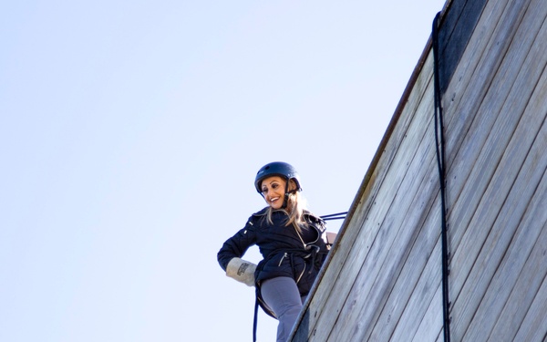 A woman wearing a blue helmet and black jacket is rappelling or abseiling down a large wooden wall structure. She is smiling and appears confident as she descends using a rope and harness system. The photo is taken from a low angle against a clear blue sky.