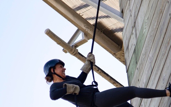 A woman is rappelling down a wooden wall structure, suspended by ropes while wearing safety gear. She is smiling and appears confident, with her feet braced against the vertical wooden surface. The photo is taken from a low angle looking upward, emphasizing the height of the activity.
