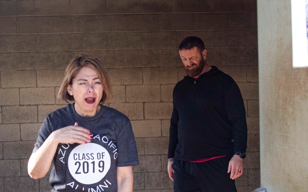Two individuals are walking through what appears to be a covered outdoor passageway or tunnel between buildings. The woman in the foreground is wearing an 'Azusa Pacific Alumni Class of 2019' t-shirt and appears to be reacting with surprise or emotion, while a bearded man in black athletic wear follows behind her. The setting appears to be a campus or institutional environment with concrete block walls.