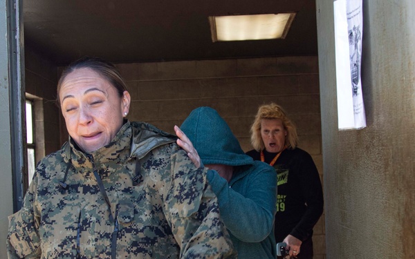 The image shows three individuals exiting or standing near a doorway of a concrete block building with graffiti on the exterior wall. A woman in U.S. Marine Corps MARPAT digital camouflage uniform appears to be guiding or escorting a civilian in a teal hoodie and colorful leggings. A third woman in a black jacket with an orange lanyard is visible in the background doorway.