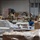 Military personnel in camouflage uniforms are working inside a warehouse facility, sorting and packing food items into cardboard boxes. The scene appears to be a food bank or emergency food distribution operation, with multiple soldiers wearing gloves handling canned goods and other food products. The warehouse contains large quantities of boxed and palletized food supplies.
