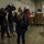 A military press conference or media briefing is taking place inside what appears to be a warehouse or logistics facility. A soldier in camouflage uniform stands facing multiple news crews equipped with professional cameras and microphones. At least two reporters, including one in a red jacket and one in a teal coat, are conducting the interview.