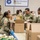 U.S. Army soldiers in OCP (Operational Camouflage Pattern) uniforms are volunteering at the Greater Cleveland Food Bank, packing cardboard boxes with food items. Multiple soldiers, both male and female, are working alongside civilian volunteers at packing tables. The scene depicts a military community service operation at a food bank facility.