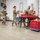 Five U.S. Army soldiers in MultiCam/OCP camouflage uniforms are working in a warehouse facility, appearing to pack or sort items into brown paper bags and red plastic crates. The setting appears to be a food bank or distribution center, with pallets of boxed goods visible in the background. The soldiers are engaged in what appears to be a humanitarian or community support mission.