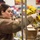 A woman in a tan fleece jacket wearing white latex gloves is handling a yellow bag of food at what appears to be a food bank or distribution center. Several individuals in military camouflage uniforms are working in the background, packing cardboard boxes. The setting suggests an organized food distribution or volunteer operation.