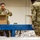Two U.S. Army soldiers in OCP (Operational Camouflage Pattern) uniforms are working at a food bank or distribution warehouse, packing cardboard boxes on a blue conveyor belt. The soldier in the foreground, a woman, is placing canned goods into a box labeled 'Shelf Stable KIT.' This appears to be a military humanitarian assistance or community support operation.