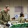 Ohio National Guard soldiers are working at what appears to be a food bank or food distribution center. The foreground soldier, identified by name tape as 'KELLEY' and wearing an Ohio state flag patch, is sorting and organizing food items while wearing black gloves. Multiple soldiers in OCP (Operational Camouflage Pattern) uniforms are visible working along the distribution tables.