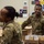 U.S. Army soldiers in OCP (Operational Camouflage Pattern) uniforms are engaged in a community service or humanitarian aid activity, packing cardboard boxes with canned goods. A smiling male soldier with the name tape 'SMITH' and 'U.S. ARMY' patch is prominently visible in the foreground. The setting appears to be an indoor facility such as a food bank or community center.