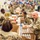 A large group of U.S. Army soldiers in OCP (Operational Camouflage Pattern) uniforms are working at a food bank or distribution warehouse, packing boxes with canned goods and other food items. The soldiers are wearing latex gloves and working along assembly-style tables in what appears to be a community service or disaster relief operation. The warehouse background features colorful t-shirts or banners displayed on the wall.