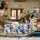 Three U.S. Army soldiers in OCP (Operational Camouflage Pattern) uniforms are working at a food bank or distribution center, packing food items into boxes labeled 'Shelf Staple KIT'. The soldiers are handling canned goods and packaged food items, suggesting a humanitarian or community support mission. The setting appears to be a large warehouse or distribution facility.