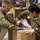 Two U.S. Army soldiers in MultiCam/OCP camouflage uniforms are working together to seal a cardboard box labeled '2' using packing tape. Both soldiers are wearing protective gloves, suggesting a health or safety-conscious operation, likely logistics or supply distribution. The background shows a warehouse or distribution center with colorful banners or flags on the wall.
