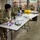 Multiple U.S. Army soldiers in OCP (Operational Camouflage Pattern) uniforms are working inside a warehouse facility associated with a 'Harvest for Hunger' food bank or distribution organization. The soldiers appear to be sorting and packaging supplies, with some wearing protective gloves. The setting suggests a humanitarian assistance or community support mission.
