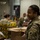 Military personnel in camouflage uniforms are working at what appears to be a food bank or disaster relief distribution center. Multiple soldiers are sorting and packing canned goods and other food items into cardboard boxes. The primary subject in the foreground is a smiling female soldier wearing latex gloves and handling a cardboard box.