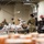 Military personnel in camouflage uniforms are working at a food bank or distribution center, sorting and packing food items into boxes and bags. The setting appears to be a warehouse facility with long tables covered in food products and supplies. A visible sign reading 'Combat Hunger' suggests this is a charitable food distribution operation.