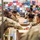 A group of military personnel in camouflage uniforms are volunteering at what appears to be a food bank or community service event, packing cardboard boxes with canned goods along an assembly line. The background wall is decorated with a colorful collage of branded t-shirts or flags from various organizations and sponsors. The scene depicts organized community service work by U.S. Army soldiers.