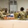 Military personnel in U.S. Army camouflage uniforms are sorting and packing canned goods at what appears to be a food bank or distribution warehouse. Several soldiers wearing protective gloves are organizing canned food items, including visible Del Monte brand products, into cardboard boxes. Additional personnel and civilians are visible working in the background of the warehouse facility.