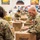 A group of U.S. military personnel in Army Combat Uniforms (ACU/OCP pattern) are volunteering at what appears to be a food bank or distribution center. Soldiers are packing cardboard boxes with food items, with a prominent 'Natrel' branded box visible in the foreground. The setting appears to be a warehouse-style facility with colorful posters and product labels on the walls.