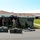 Military personnel in camouflage uniforms are assembling a large dark-colored field tent or shelter structure on a grass lawn adjacent to what appears to be a medical facility, indicated by an 'Emergency/Urgencias' sign visible in the background. Several hard-shell equipment cases are staged on the pavement in the foreground. The scene suggests a field medical or operational setup, possibly in response to a disaster or emergency situation.