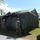 Military personnel in camouflage uniforms are erecting or assembling a large olive-drab military field tent on a grass area. The setting appears to be a military installation or base, identifiable by the Spanish-style architecture in the background and palm trees. Several service members wearing hard hats are actively working together to raise and secure the tent structure.