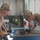 Two military personnel in tan/olive uniforms are working together at a metal workbench in what appears to be a military workshop or fabrication facility. Both individuals are wearing tan hard hats with insignia, safety glasses, and black work gloves. They appear to be assembling or inspecting a metal component on the workbench.