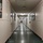 A long, empty institutional corridor with white paneled walls and a dark tiled floor receding into the distance. The hallway is lined with multiple doors on both sides, framed pictures/posters on the walls, and fluorescent ceiling lights. The setting appears to be a school, dormitory, or office building based on the architectural style and wall decorations.