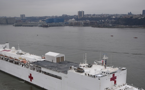 A large white hospital ship marked with prominent red crosses is navigating a river, assisted by tugboats. The name 'COMFORT' is visible on the bow of the vessel, identifying it as the USNS Comfort, a US Navy hospital ship. The ship is surrounded by tugboats and is passing by an urban waterfront skyline in overcast conditions.