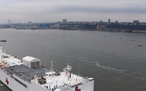 A large white US Navy hospital ship, the USNS Comfort (T-AH-20), is navigating a river waterway accompanied by a tugboat. The vessel is prominently marked with large red cross symbols on its hull and superstructure, identifying it as a medical ship. A urban skyline with buildings and trees is visible in the background under an overcast sky.
