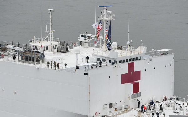 The image shows the U.S. Naval Hospital Ship COMFORT, a large white vessel marked with prominent red crosses indicating its medical/humanitarian role. A red tugboat is visible alongside the ship, assisting with navigation. Personnel are visible on multiple deck levels of the hospital ship.