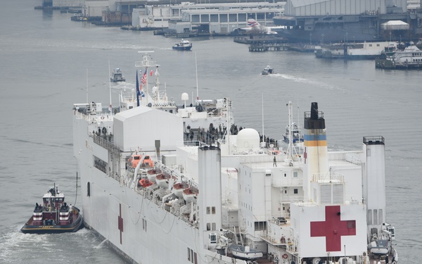 The image shows the USNS Comfort (T-AH-20), a U.S. Naval Hospital Ship, being escorted by multiple tugboats through a harbor. The large white vessel is clearly marked with red crosses and the text 'U.S. NAVAL HOSPITAL SHIP COMFORT' on its hull. The scene appears to be in an urban port environment, likely New York Harbor, with industrial dock facilities visible in the background.