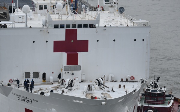 The image shows the U.S. Naval Hospital Ship COMFORT, a large white vessel prominently marked with multiple red cross symbols indicating its medical/humanitarian role. The ship is docked or being maneuvered with the assistance of tugboats on either side. Several personnel are visible on the deck, and the ship appears to be in a port or harbor setting under overcast conditions.