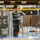 A female military service member in OCP (Operational Camouflage Pattern) uniform is working in a warehouse or food distribution facility. She is wearing gloves and handling cardboard boxes placed on a metal wire utility cart. The background shows shelving units stocked with canned goods and palletized supplies.