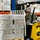 A person wearing military camouflage uniform operates a yellow Yale forklift inside a large warehouse. The forklift is carrying a large pallet stacked with what appears to be cases of bottled water wrapped in plastic. The warehouse contains extensive shelving with additional palletized goods stored on orange and blue industrial racking.